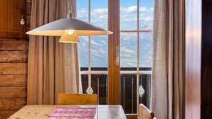 En stuga med träpanel, ett bord och en stol, en hängande lampa och ett stort fönster med utsikt över bergen.