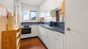 Ett kompakt kök med vita skåp, en inbyggd ugn, en diskmaskin och ett fönster som släpper in naturligt ljus.