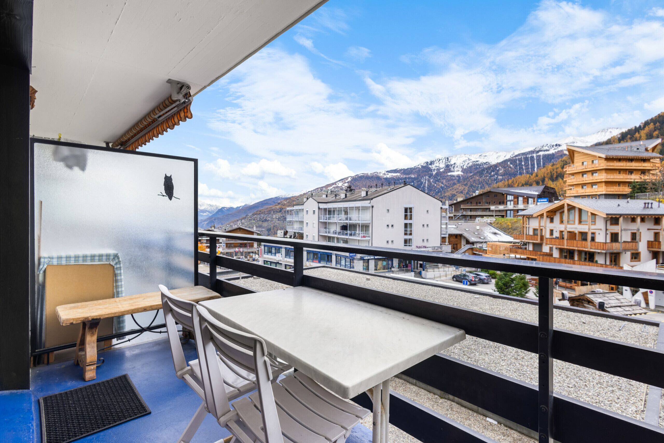 A balcony with a table and chairs, overlooking a town with buildings and mountains.