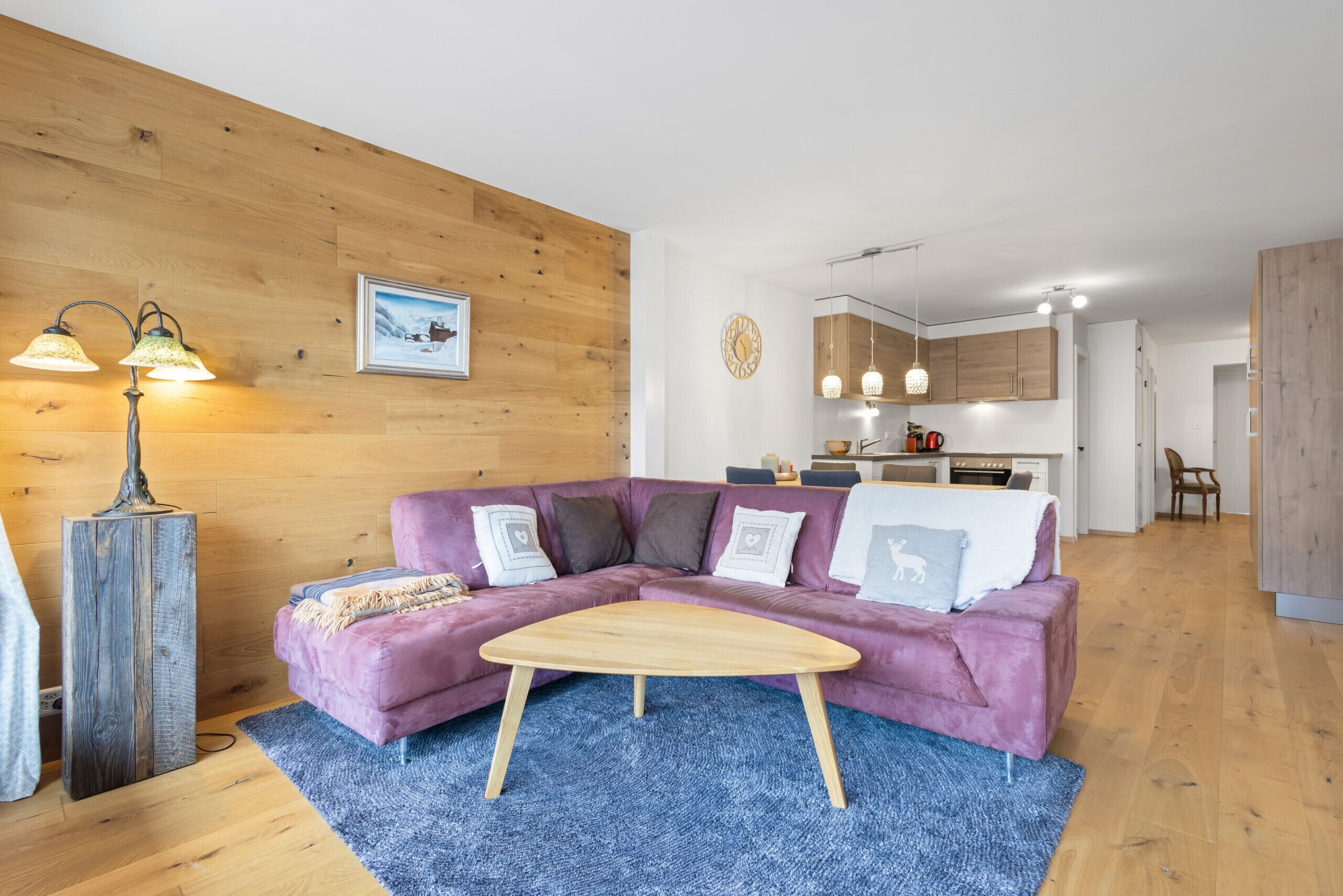 A modern living room with a wooden accent wall, a purple sofa, a wooden coffee table, and a kitchen in the background.