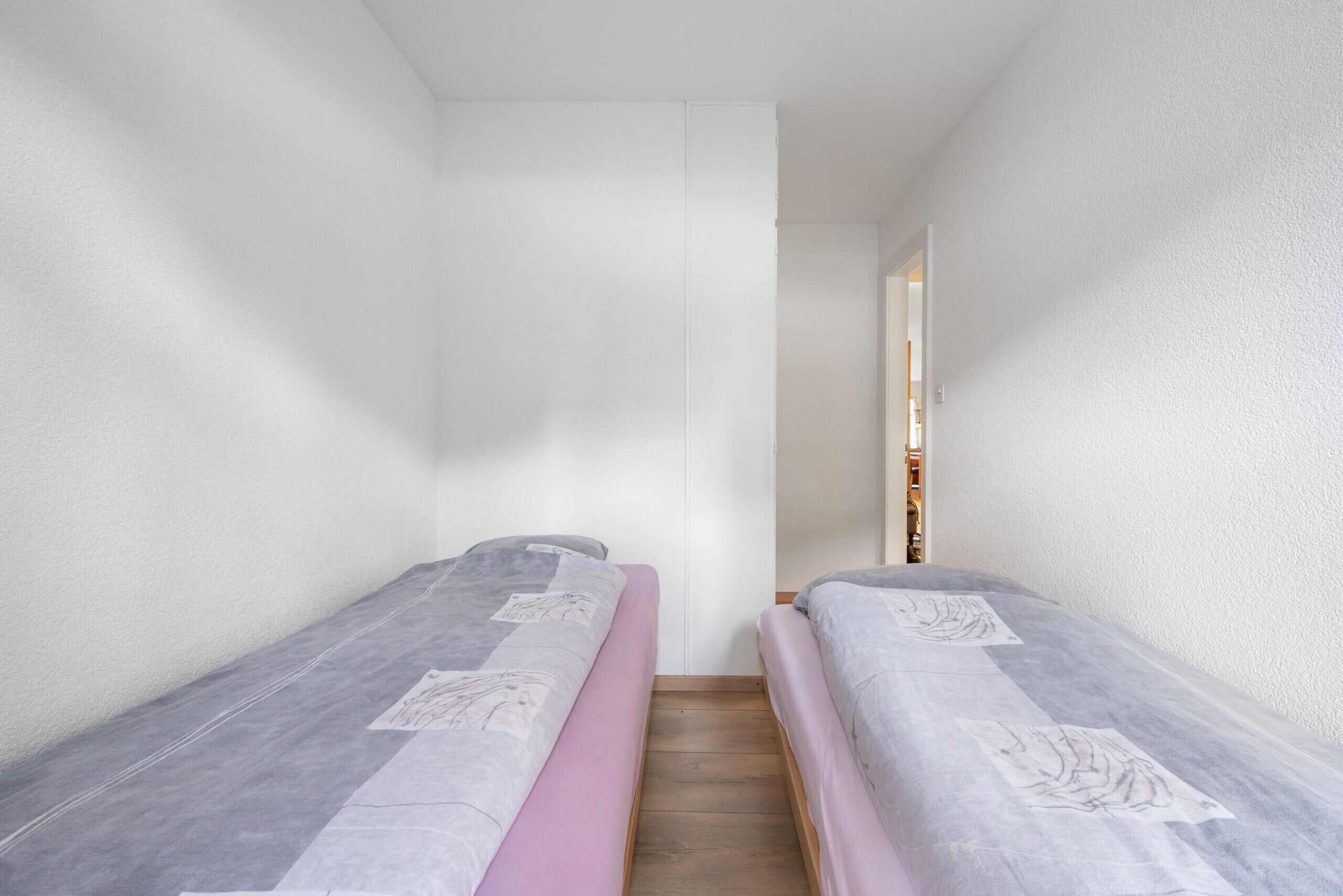 A small, narrow room with two single beds, each with a mattress and a blanket. The room has white walls and a wooden floor.