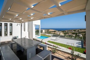 A modern outdoor patio with a pool, clear blue sky, and a view of the sea. - 3 Bedroom Villa Chara (Agia Pelagia)