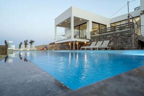 A modern poolside area with a clear view of the pool, lounge chairs, and a contemporary building with large windows. - 3 Bedroom Villa Chara (Agia Pelagia)