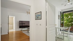 A modern living room with a flat-screen TV, a dining area with a glass table and chairs, and a large window with a view of greenery. - Elegant New Apartment With Balcony in the 16th - M (Paris)