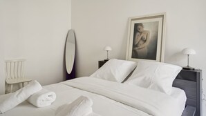 A neatly made bed with white linens, two bedside lamps, a framed picture on the wall, and a white chair. - Elegant New Apartment With Balcony in the 16th - M (Paris)
