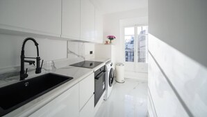 Une cuisine moderne avec des meubles blancs, un robinet noir, un four encastré et une fenêtre donnant sur des immeubles.