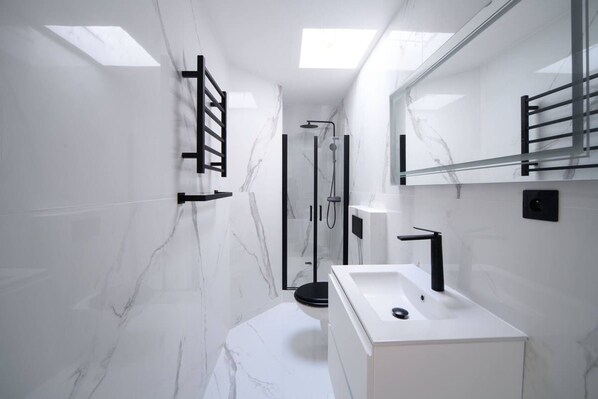 Un baño moderno con pared de mármol blanco, ducha envuelta en vidrio y lavamanos blanco con grifo negro.