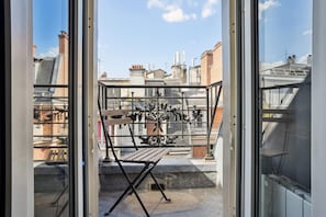 On aperçoit, à travers une porte vitrée, un balcon doté d’une rambarde en métal et d’un banc en bois.