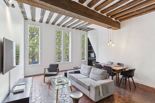 A modern living room with a sofa, chairs, a glass coffee table, and a dining area with a table and chairs. Large windows provide natural light and views of greenery outside. - Exceptional Appt - Saint-germain- Mobility Lease (Paris)