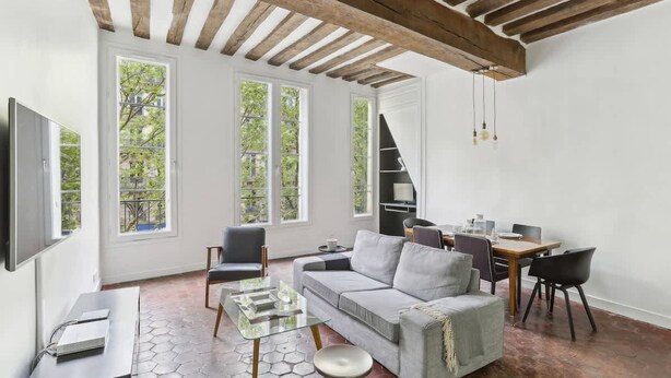 A modern living room with a grey sofa, a glass coffee table, and a dining area with a wooden table and chairs. Large windows provide natural light and views of greenery outside. - Exceptional Appt - Saint-germain- Mobility Lease (Paris)