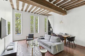 A modern living room with a grey sofa, a glass coffee table, and a dining area with a wooden table and chairs. Large windows provide natural light and views of greenery outside. - Exceptional Appt - Saint-germain- Mobility Lease (Paris)