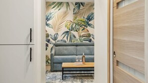 Ett rum med en grå soffa, ett träbord med glas och en flaska, och en vägg med ett tropiskt bladmotiv.