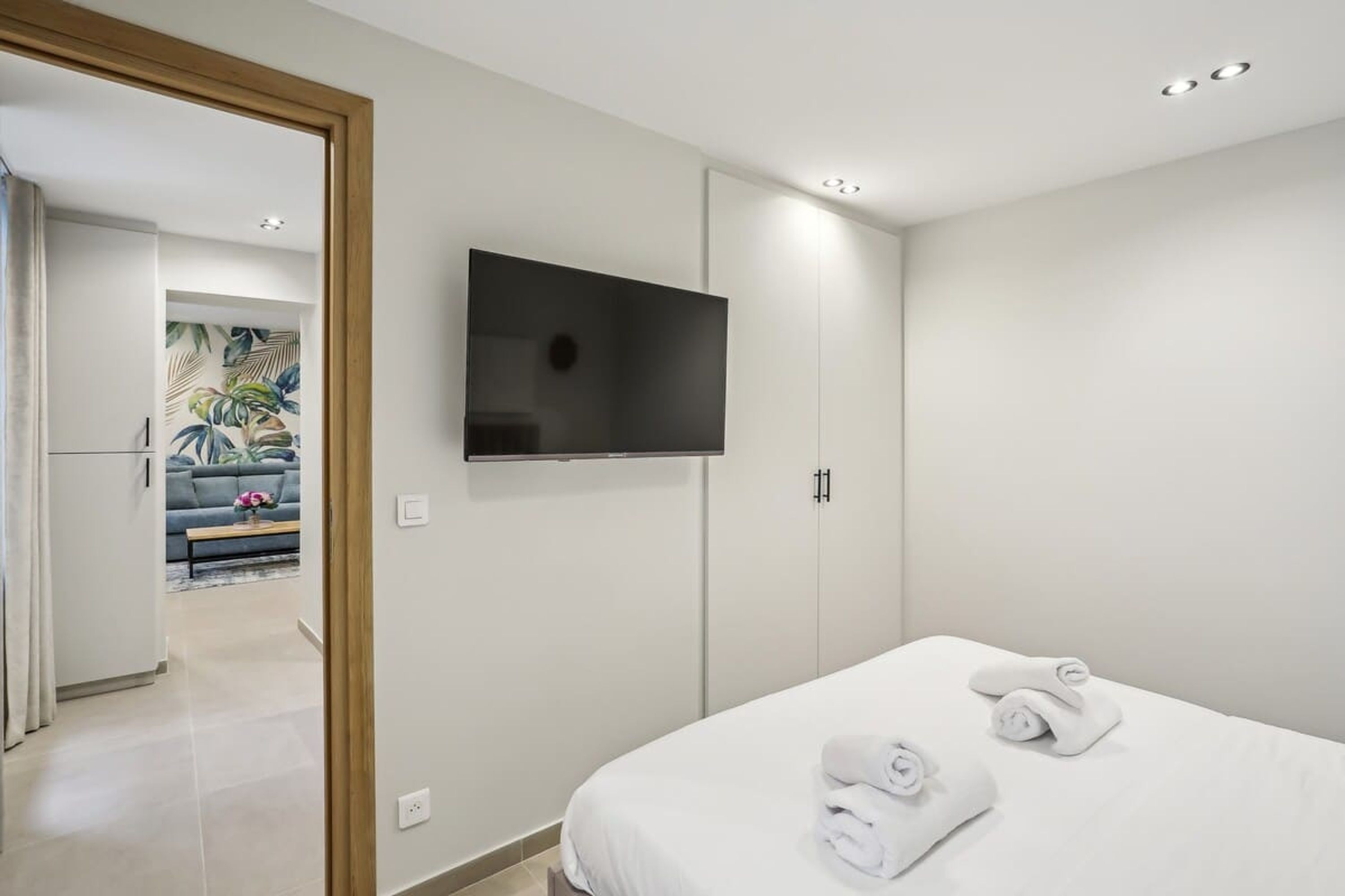 A modern hotel room with a bed, a flat-screen TV mounted on the wall, and a view into the living area.