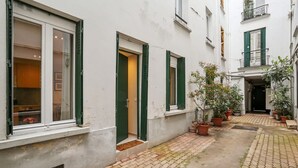 En smal gränd med en vit byggnad, gröna fönsterluckor och krukväxter.