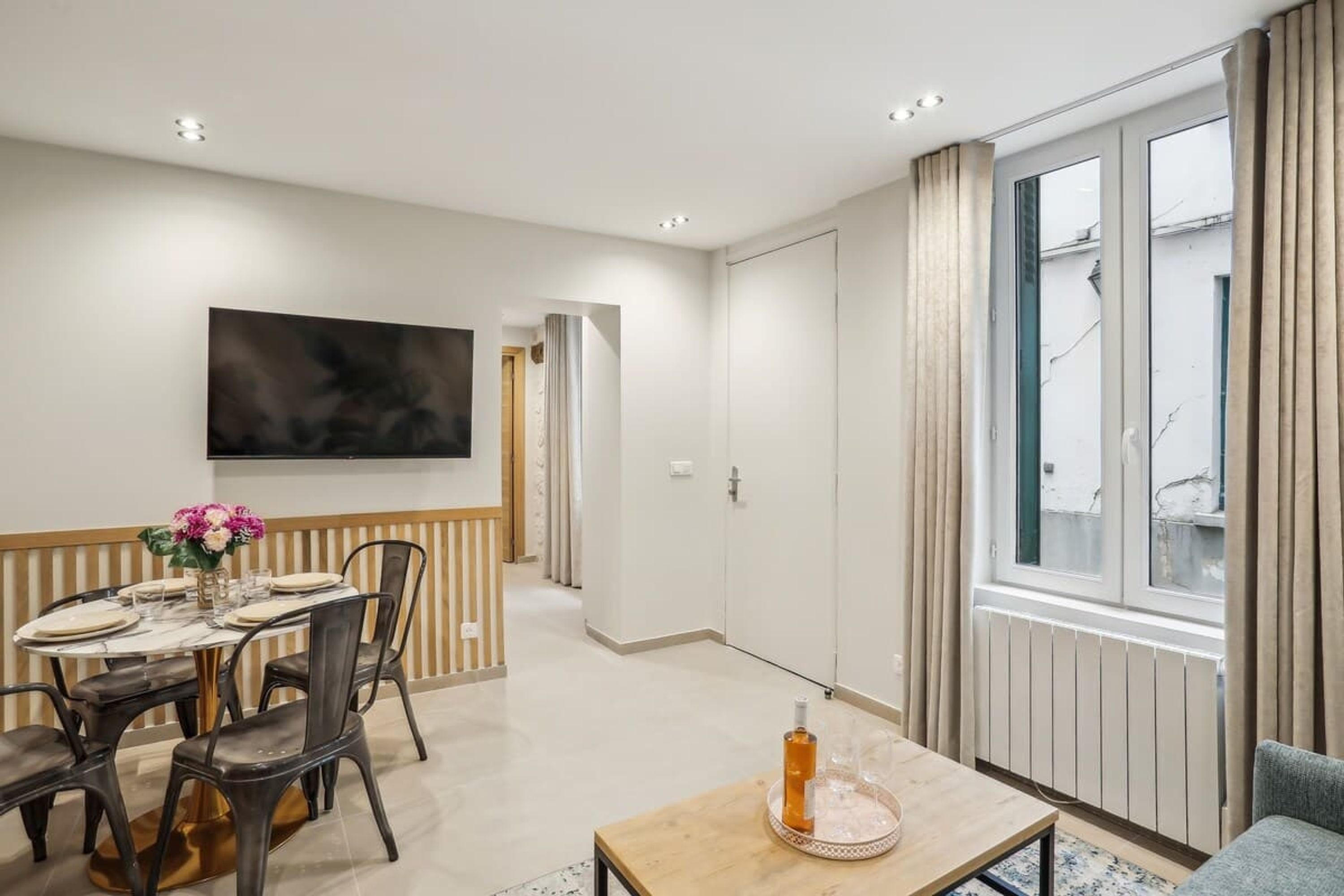 Un salon moderne comprenant un coin repas, un téléviseur à écran plat fixé au mur et une grande fenêtre avec des rideaux.