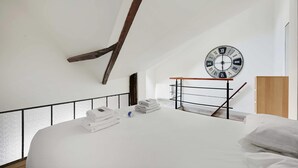 A modern hotel room with a large bed, neatly folded white towels, a wooden headboard, a wall-mounted clock, and a small table with a metal frame. - Authentic Apartment - 1BR 6P - Centre de Paris (Paris)