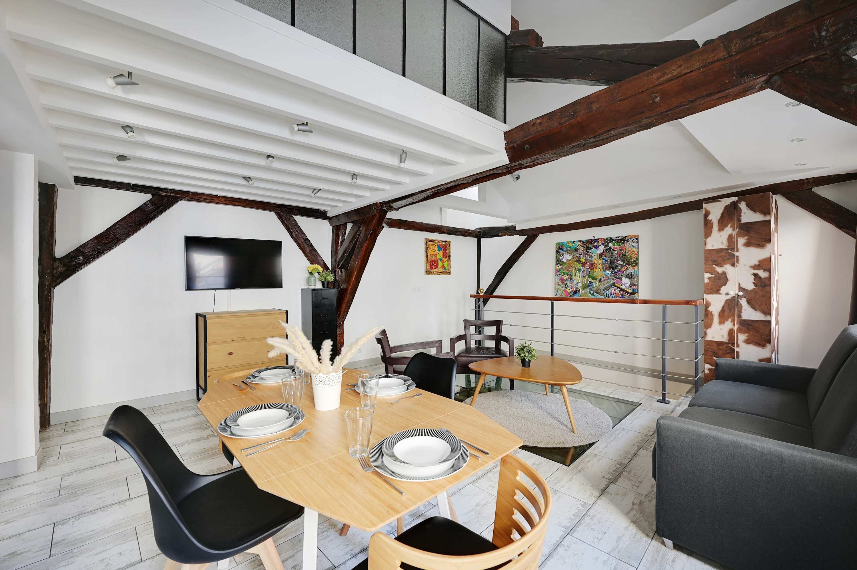 Ein moderner Essbereich mit einem Holztisch, gedeckt für vier Personen, umgeben von schwarzen Stühlen. Es gibt einen kleinen runden Tisch mit einem einzelnen Stuhl und eine Couch im Hintergrund. Der Raum zeichnet sich durch freiliegende Holzbalken und eine weiße Decke aus.