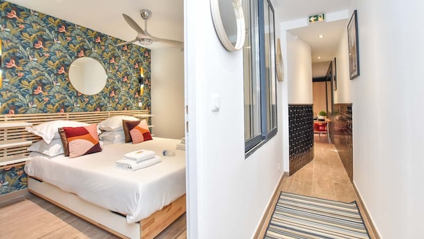 Une chambre d’hôtel avec un lit, un ventilateur de plafond, un mur décoré d’un motif, une fenêtre et un couloir menant à un espace repas.