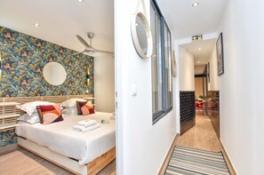 Une chambre d’hôtel avec un lit, un ventilateur de plafond, un mur décoré d’un motif, une fenêtre et un couloir menant à un espace repas.