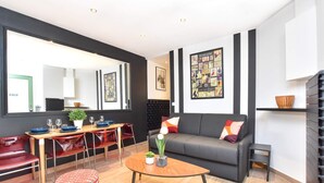 Un salon moderne avec un canapé, une table à manger et des chaises. On y trouve un grand miroir, des œuvres d’art accrochées aux murs et une petite table d’appoint.