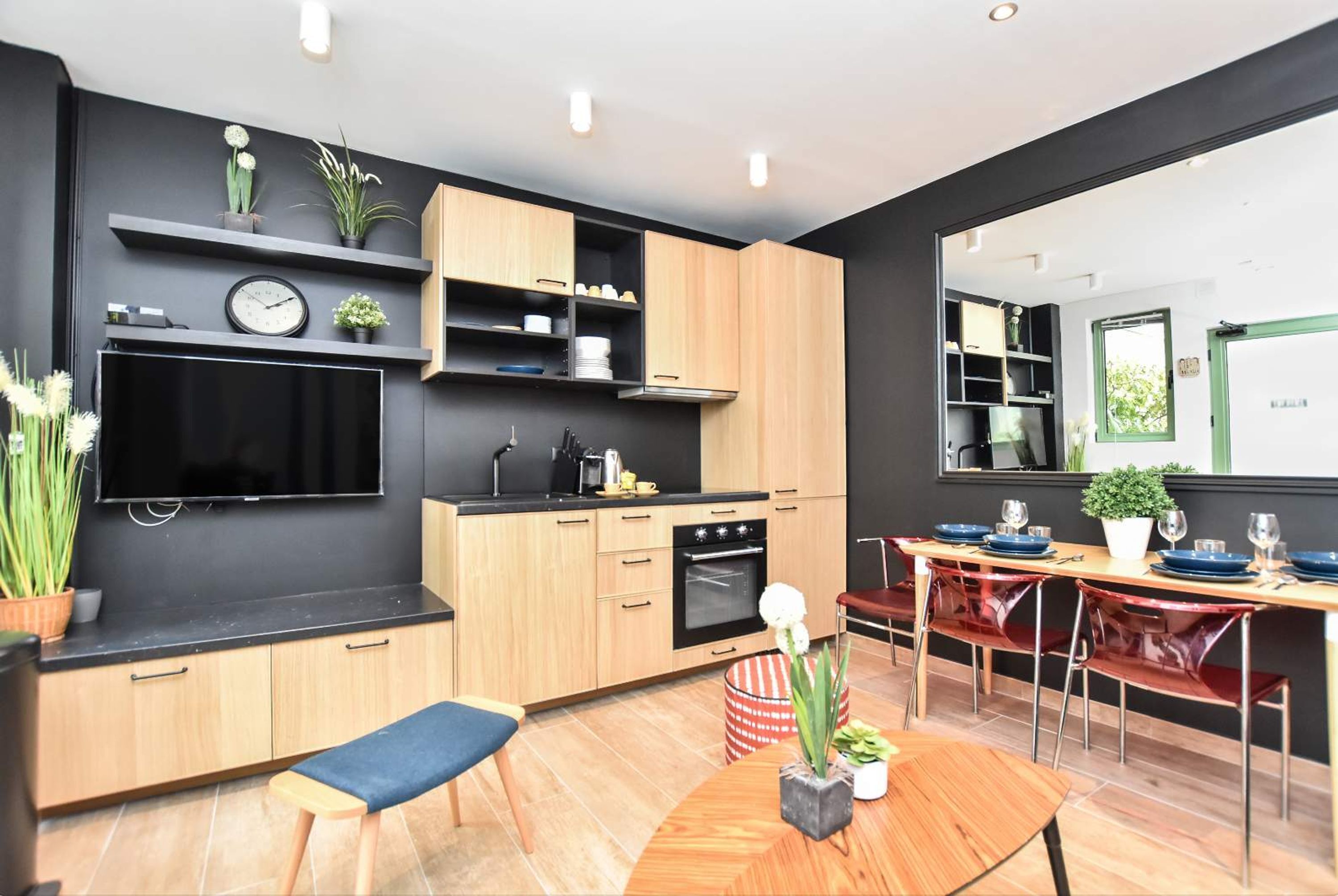 A modern kitchen with wooden cabinets, a black countertop, and a built-in oven. There is a dining area with a wooden table and red chairs.
