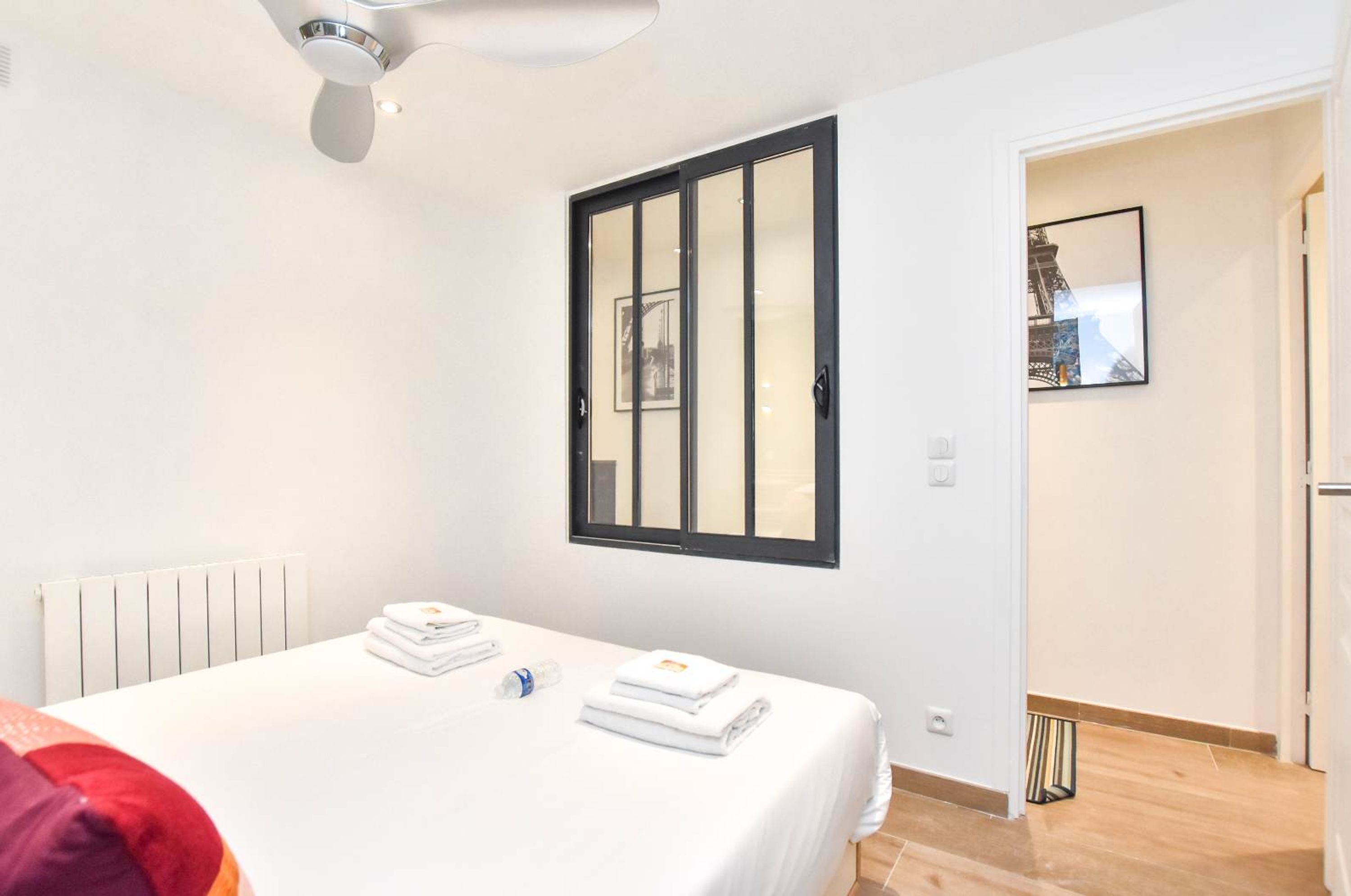 Una habitación de hotel con una cama, un radiador, un ventilador de techo, un ventanal con marco negro y una puerta que da a otra estancia.
