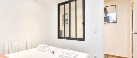 Una habitación de hotel con una cama, un radiador, un ventilador de techo, un ventanal con marco negro y una puerta que da a otra estancia.