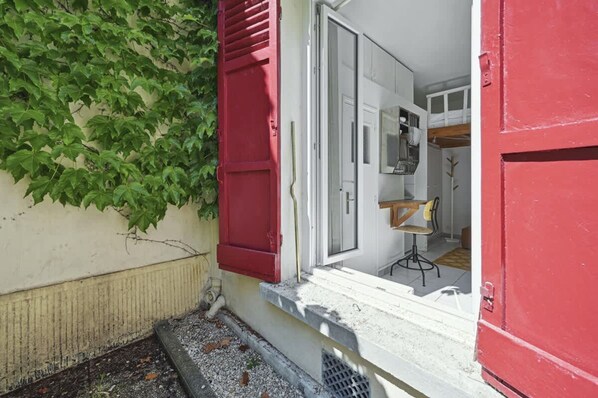Une entrée étroite, avec une porte rouge et un petit seuil, mène à un espace intérieur blanc où se trouvent une chaise et une table.