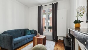 A modern living room with a blue sofa, a wooden coffee table, and a small round ottoman. A white rug is on the wooden floor, and there is a window with curtains. - Warm Apartment - Montmartre - Mobility Lease (Paris)