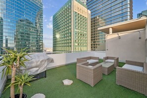 Eine Dachterrasse mit Kunstgrasanlage, Korbmöbeln und Blick auf hohe Gebäude.