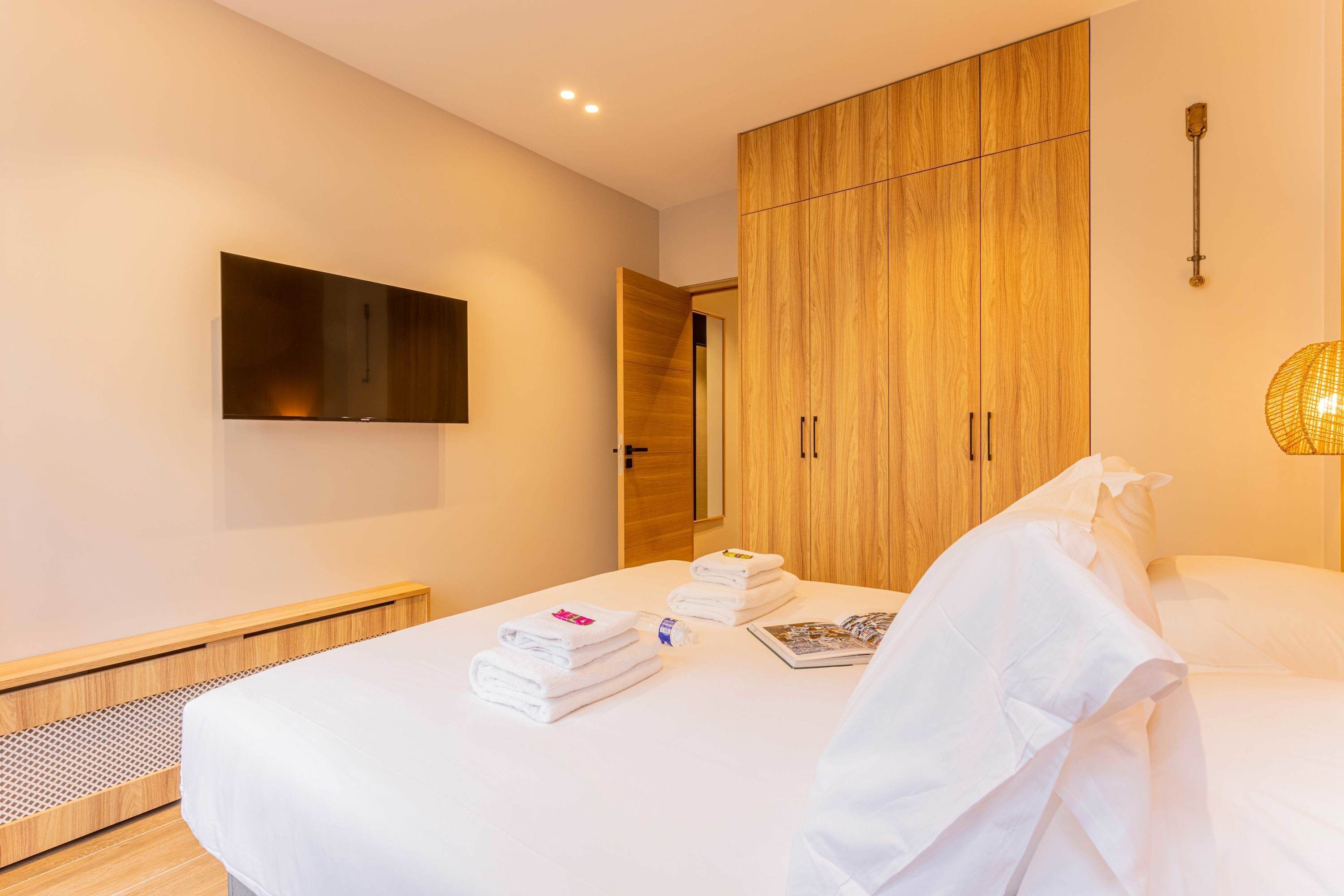 A hotel room with a bed, a TV mounted on the wall, wooden wardrobe, and a bedside lamp.