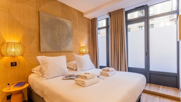 Quarto de hotel com uma cama grande, duas luminárias de cabeceira, uma obra de arte na parede e vista através de uma porta de vidro.