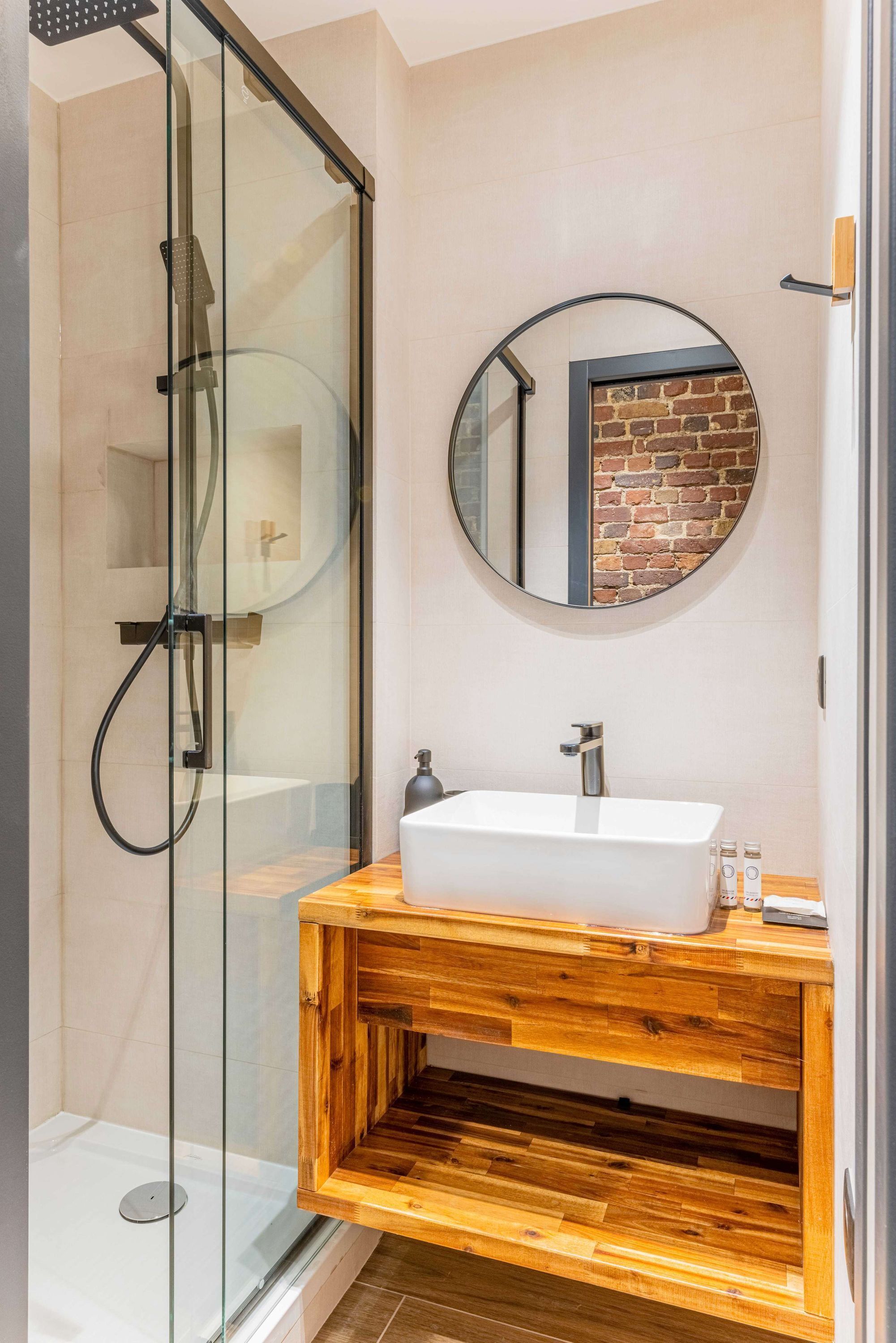 A modern bathroom with a glass-enclosed shower, a wooden vanity with a white sink, and a round mirror above it.