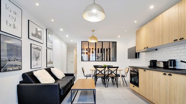 Eine moderne Küche mit Holzausstattung, ein Essbereich mit schwarzem Sofa und Couchtisch, sowie eine Wand mit gerahmten Bildern.