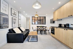 A modern kitchen with wooden cabinets, a dining area with a black sofa and a coffee table, and a wall with framed pictures. - Super Appartment-2br 6p-bastille (Paris)