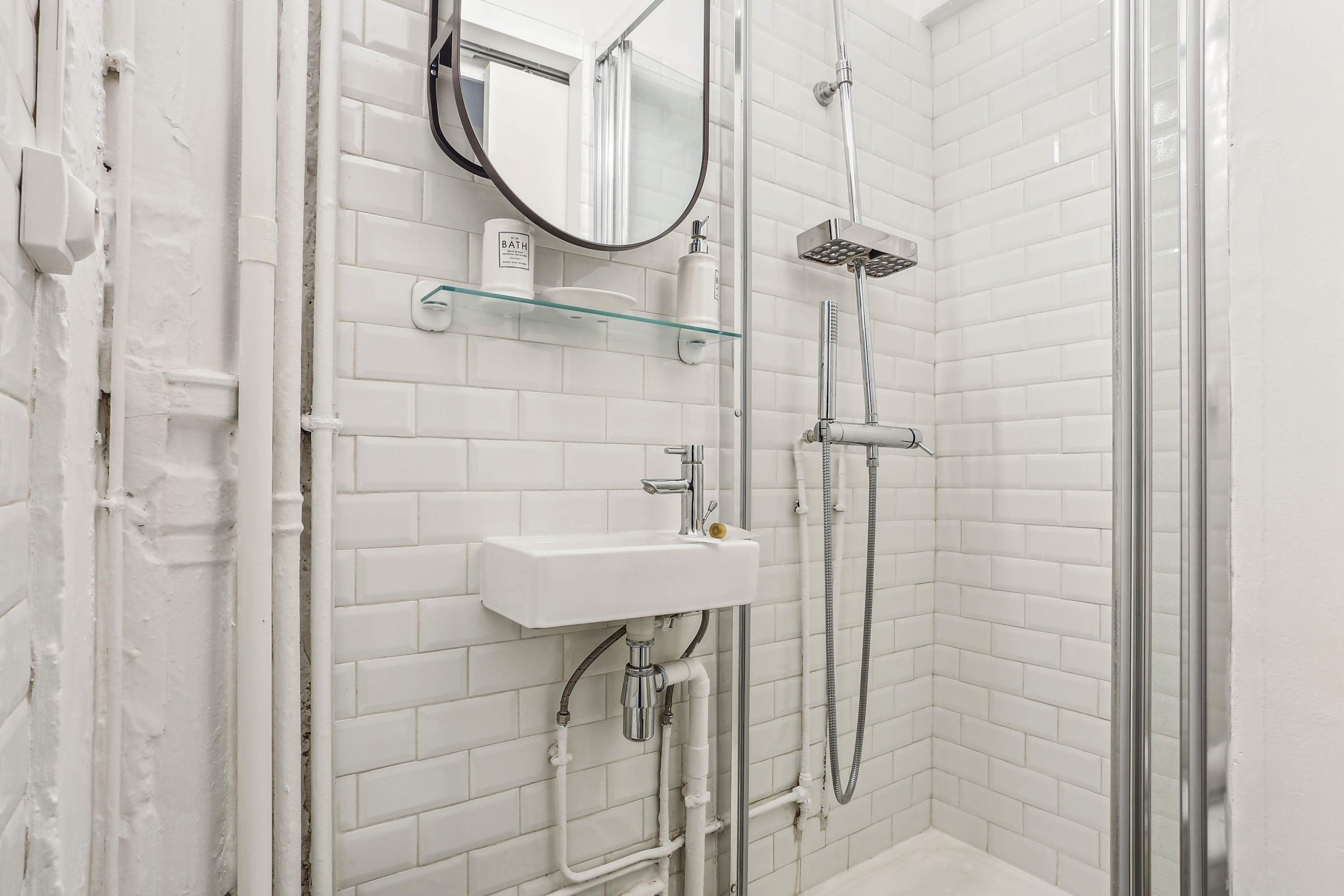 A modern bathroom with white tiled walls, a glass shower enclosure, a white sink with a mirror above it, and a glass shelf with toiletries.