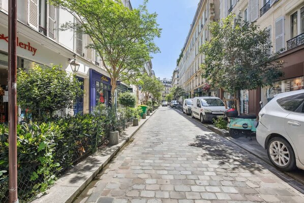 Une ruelle étroite pavée, bordée d’immeubles, de voitures garées et d’une végétation luxuriante.