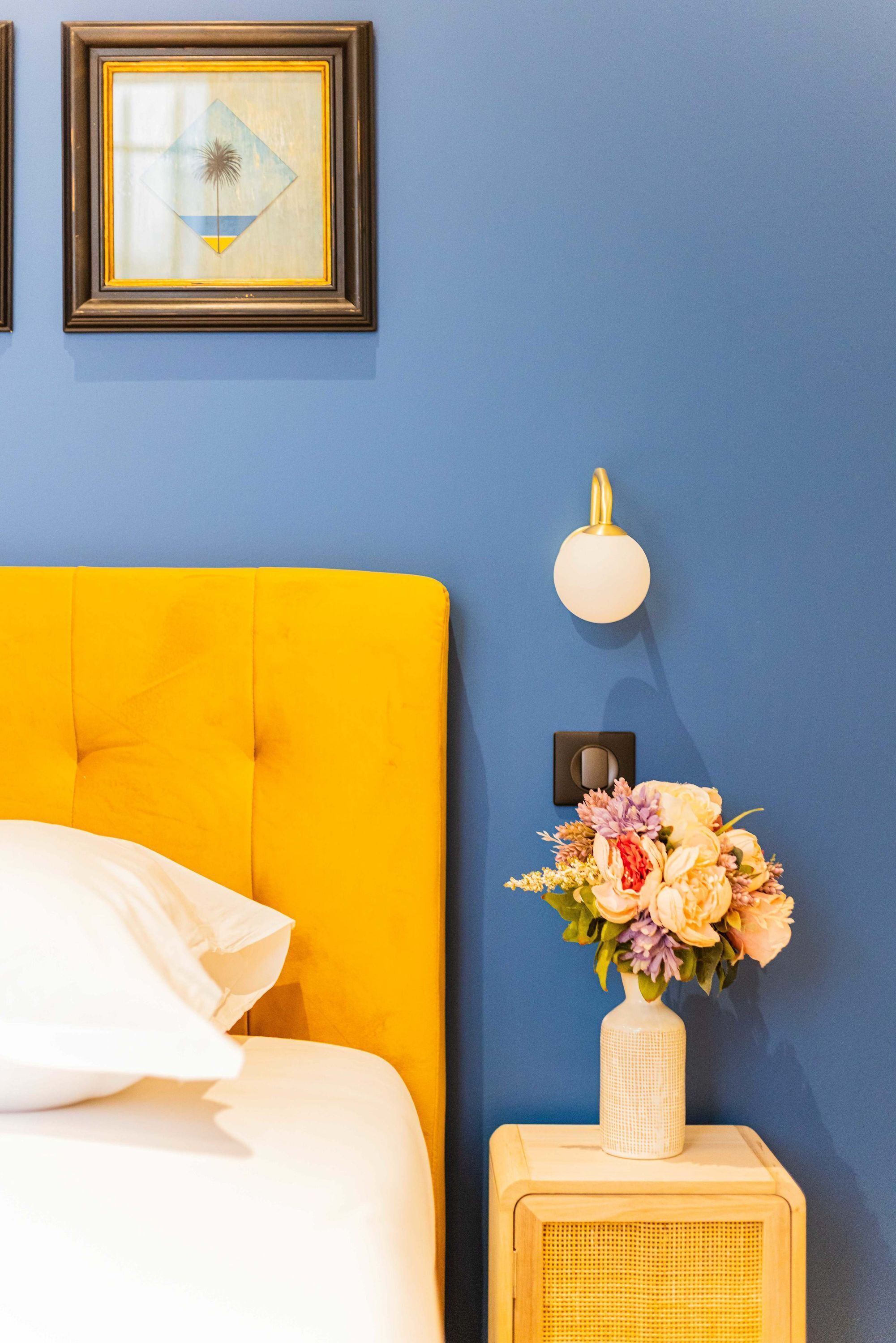 A bed with a yellow headboard, a white pillow, a wall-mounted light fixture, and a vase with flowers on a bedside table.