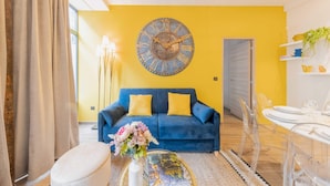 A living room with a blue sofa, a round wall clock, and a dining area with clear chairs. - Amazing Apartment - 2BR 6P - Gare du Nord (Paris)