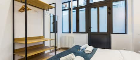 A bedroom with a bed, a wooden shelf unit, and a large window with a view of buildings.