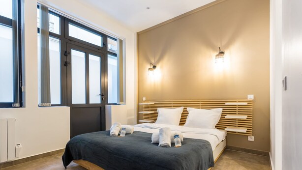 A modern hotel room with a large bed, two bedside lamps, a window with blinds, and a radiator. - Amazing Apartment - 2br6p - Bastille (Paris)