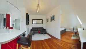 A modern living room with a sofa, a dining table, a kitchen area with a red cabinet, and a large mirror. - A Cozy Refuge - 18th - Mobility Lease (Paris)