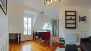 A modern living room with a wooden floor, a black sofa, a dining area with a red table, and a kitchen with a window. - A Cozy Refuge - 18th - Mobility Lease (Paris)