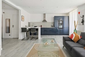 A modern living room with a glass coffee table, a grey sofa, and a kitchen in the background. - Charmant Appt 1brd 2P - Neuilly-sur-seine (Neuilly-sur-Seine)