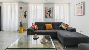 A modern living room with a grey sectional sofa, a glass coffee table, and two wall art pieces. - Charmant Appt 1brd 2P - Neuilly-sur-seine (Neuilly-sur-Seine)