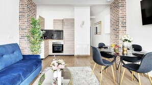 Een moderne woonkamer met een blauwe bank, een eettafel met stoelen en een keuken met een bakstenen muur en witte kasten.