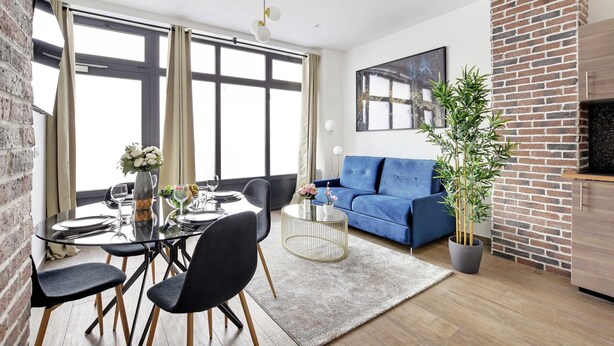 Een moderne woonkamer met een blauwe bank, een eettafel gedekt voor vier personen en een groot raam met gordijnen.