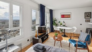 A modern living room with a city view, a dining area with a white table and chairs, and a cozy seating area with a blue chair. - Extra Studio on the 23rd Floor - Stunning View (Paris)