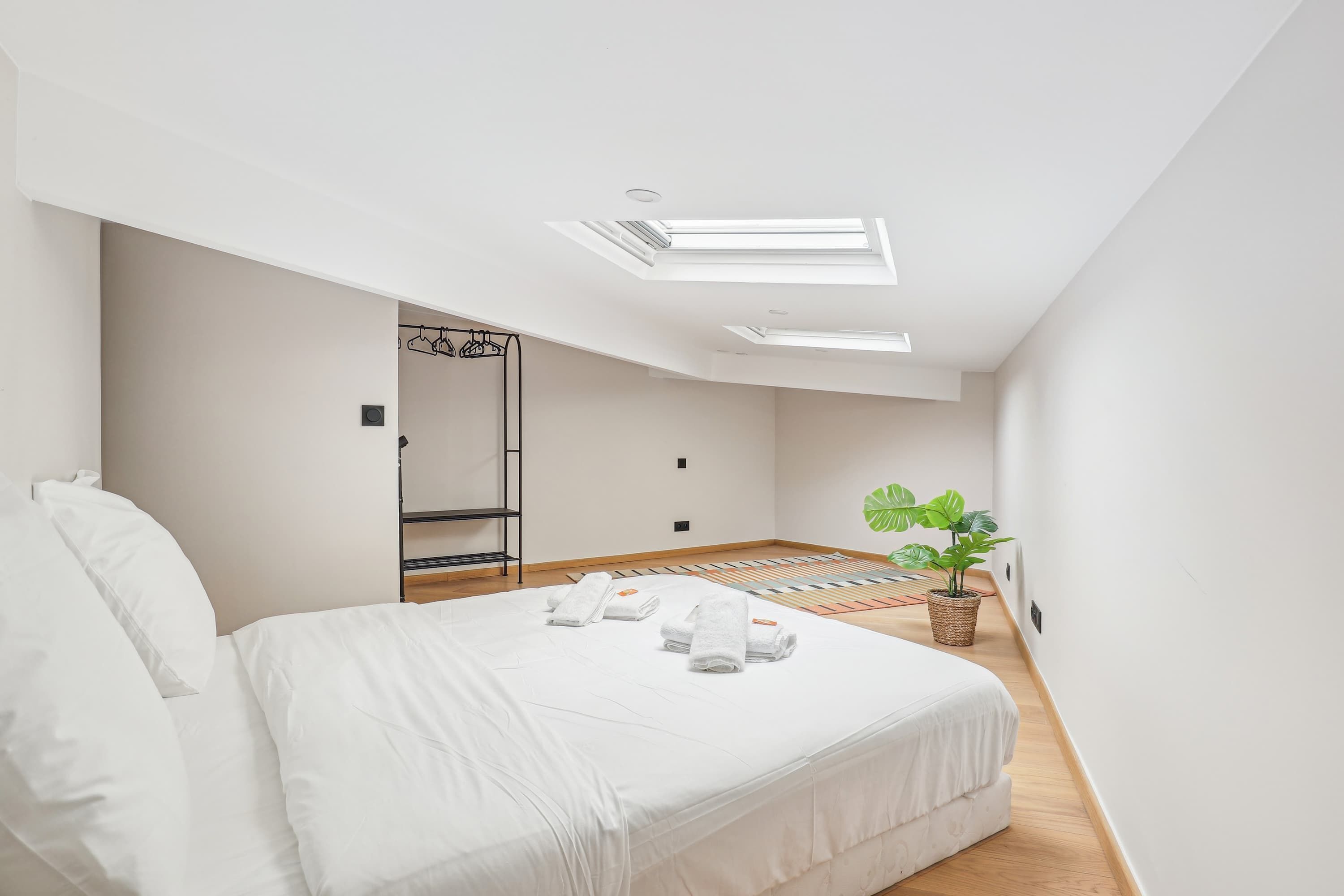 A modern bedroom with a large bed, skylights, a small plant, and a wooden floor.