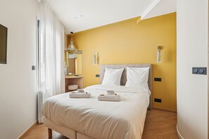 A hotel room with a bed, white linens, a yellow accent wall, a bedside table with a lamp, and a window with white curtains. - Maison Atypique de 82 m (Vincennes)